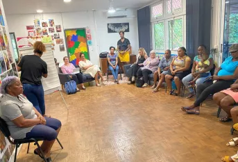 Des femmes, dont l’une tient un enfant sur ses genoux, sont assises en cercle dans une salle colorée de la Maison des Familles de Cayenne. Elles participent à un atelier sur les droits de l’enfant animé par deux intervenantes. Une femme est debout au fond de la pièce, entre deux femmes assises. A gauche, une intervenante a le dos tourné et écrit sur un paper boad.