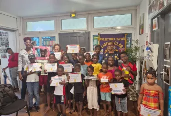 Photo d’un groupe d’enfants et d’adultes souriants dans une salle, tenant des diplômes de fin d’année qui récompensent leur assiduité à l’atelier Vivre l’école de l’année scolaire 2024-2025. Une banderole du Lions Club MFA est visible en arrière-plan. Ambiance festive et valorisante.