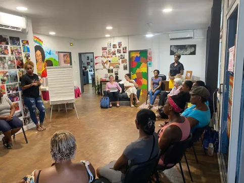 Des femmes sont assises en cercle dans une salle colorée de la Maison des Familles de Cayenne. Elles participent à un atelier sur les droits de l’enfant. Une femme est debout au fond de la pièce, entre deux femmes assises. A gauche, une intervenante est debout près d’un paper boad.