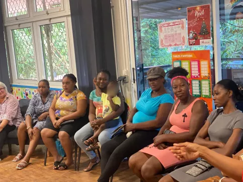 Des femmes, dont l'une a un enfant sur ses genoux, sont assises en demi-cercle dans une salle lumineuse et colorée de la Maison des Familles de Cayenne. Elles participent à un atelier sur les droits de l’enfant.  
