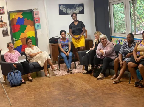 Sept femmes sont assises en demi-cercle dans une salle lumineuse et colorée de la Maison des Familles de Cayenne. Une femme est debout entre deux femmes assises. Elles participent à un atelier sur les droits de l’enfant. 