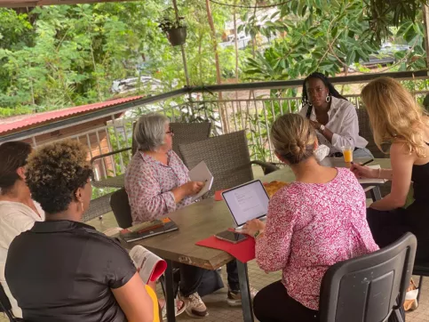 Cinq femmes, assises autour d’une table dans un espace ouvert et aéré à l’étage de la Maison des Familles de Cayenne, tournent la tête vers une sixième femme qui est assise en bout de table. Elles l’écoutent attentivement. Ces femmes participent à un atelier sur les droits de l’enfant. 