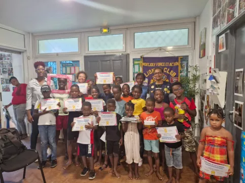 Photo d’un groupe d’enfants et d’adultes souriants dans une salle, tenant des diplômes de fin d’année qui récompensent leur assiduité à l’atelier Vivre l’école de l’année scolaire 2024-2025. Une banderole du Lions Club MFA est visible en arrière-plan. Ambiance festive et valorisante.