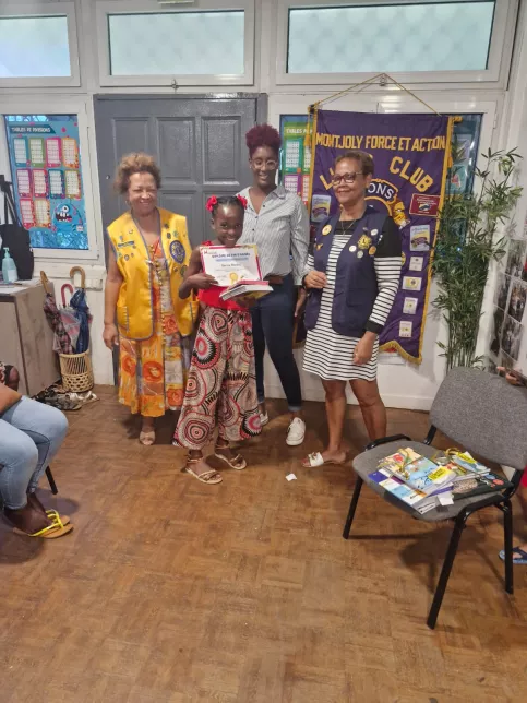 Une enfant souriante reçoit un diplôme et des livres en présence de trois femmes adultes, dont deux membres du Lions Club. Une banderole du club est accrochée au mur en arrière-plan. Ambiance chaleureuse de remise de récompense.