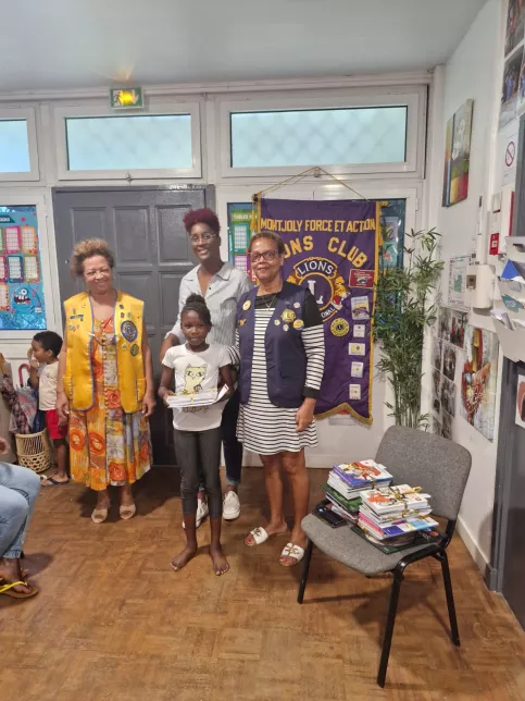 Une jeune fille tenant des livres se tient debout devant une banderole du Lions Club, accompagnée de trois femmes adultes souriantes. Une pile d’ouvrages éducatifs est visible sur une chaise à côté. Scène de remise de récompense dans un cadre convivial.