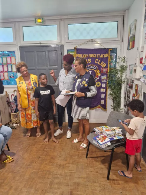 Un jeune garçon est félicité par trois femmes adultes devant une banderole du Lions Club. Une pile de livres éducatifs est posée sur une chaise, pendant qu’un petit garçon à droite photographie la scène avec un téléphone.