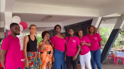 1 homme portant un tee-shirt rose fushia avec le logo de la Maison des Familles et 6 femmes, dont 4 portent aussi un tee-shirt rose fushia avec le même logo, des équipes des Maisons des Familles réunis et souriants sous un préau décoré de ballons, lors d’un moment convivial