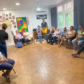 Des femmes, dont l’une tient un enfant sur ses genoux, sont assises en cercle dans une salle colorée de la Maison des Familles de Cayenne. Elles participent à un atelier sur les droits de l’enfant animé par deux intervenantes. Une femme est debout au fond de la pièce, entre deux femmes assises. A gauche, une intervenante a le dos tourné et écrit sur un paper boad.