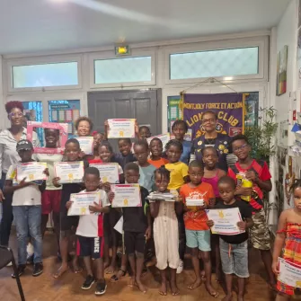 Photo d’un groupe d’enfants et d’adultes souriants dans une salle, tenant des diplômes de fin d’année qui récompensent leur assiduité à l’atelier Vivre l’école de l’année scolaire 2024-2025. Une banderole du Lions Club MFA est visible en arrière-plan. Ambiance festive et valorisante.