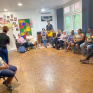Des femmes, dont l’une tient un enfant sur ses genoux, sont assises en cercle dans une salle colorée de la Maison des Familles de Cayenne. Elles participent à un atelier sur les droits de l’enfant animé par deux intervenantes. Une femme est debout au fond de la pièce, entre deux femmes assises. A gauche, une intervenante a le dos tourné et écrit sur un paper boad.