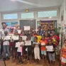 Photo d’un groupe d’enfants et d’adultes souriants dans une salle, tenant des diplômes de fin d’année qui récompensent leur assiduité à l’atelier Vivre l’école de l’année scolaire 2024-2025. Une banderole du Lions Club MFA est visible en arrière-plan. Ambiance festive et valorisante.